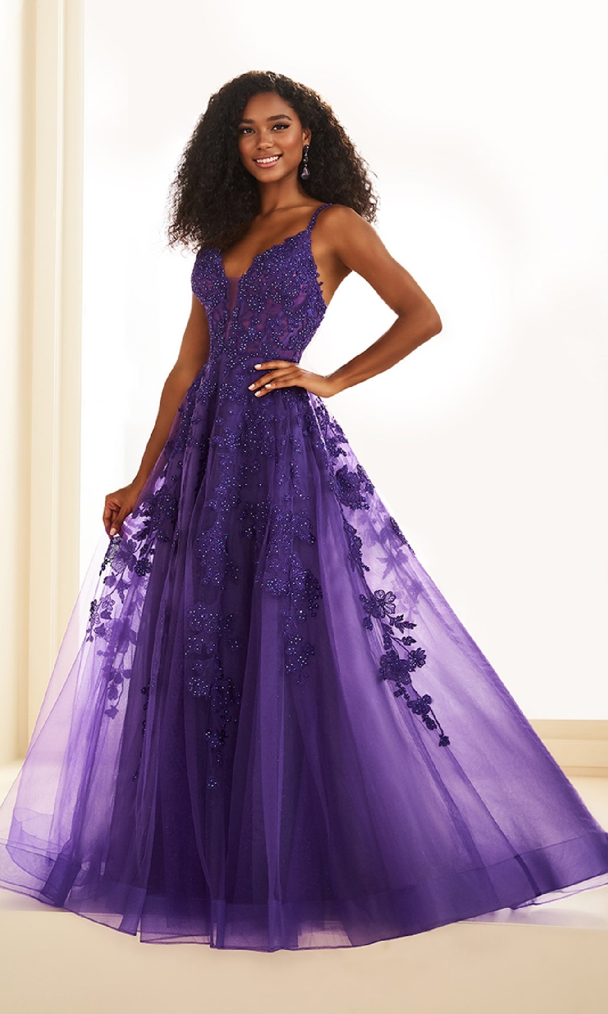 A woman smiles at the camera wearing the Ellie Wilde EW36015, a floor-length, sleeveless purple glitter tulle prom dress with lace embroidery and a sheer overlay.